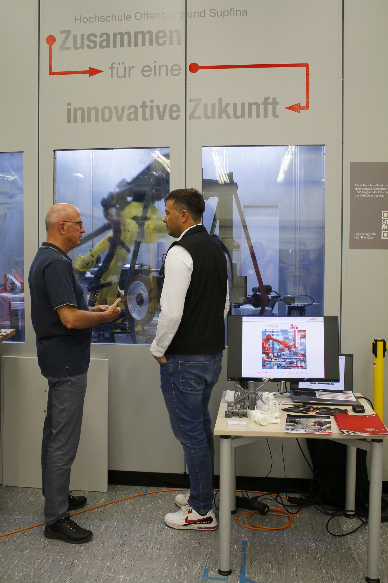 Zwei Mäner stehen vor einem Fenster, dahinter arbeitet ein Industrieroboter