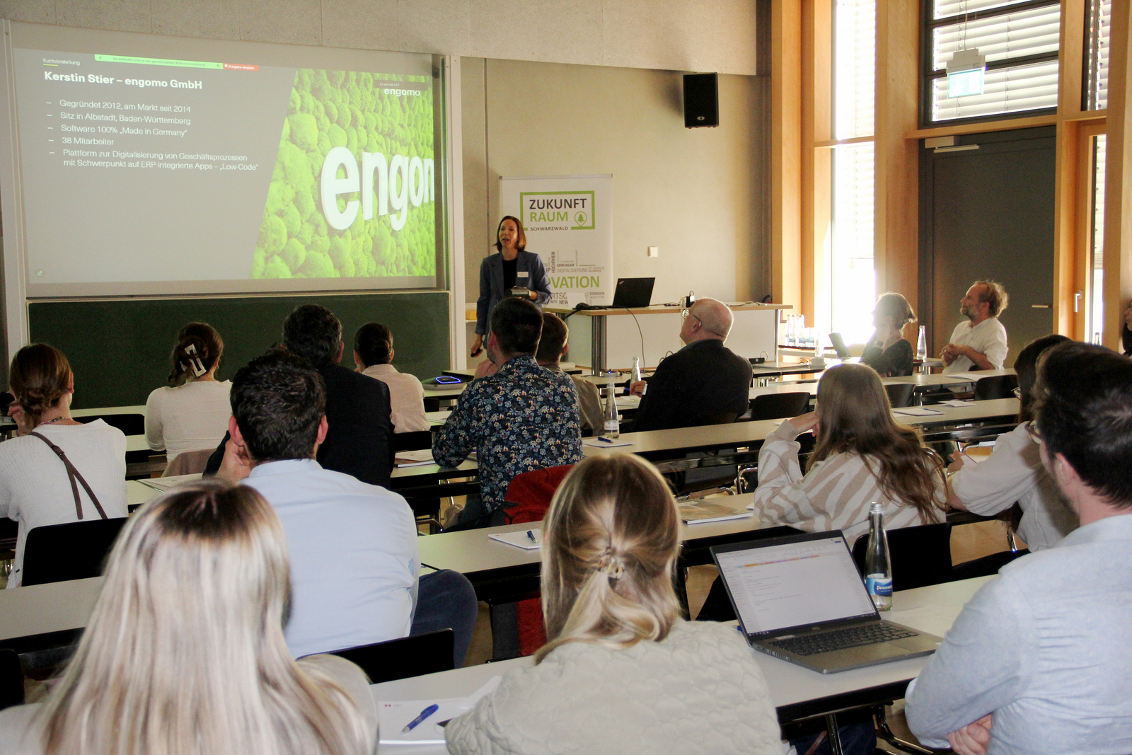 Eine Frau steht rechts einer Leinwand vor dem sitzenden Publikum und erläutert eine Präsentation auf der Leinwand