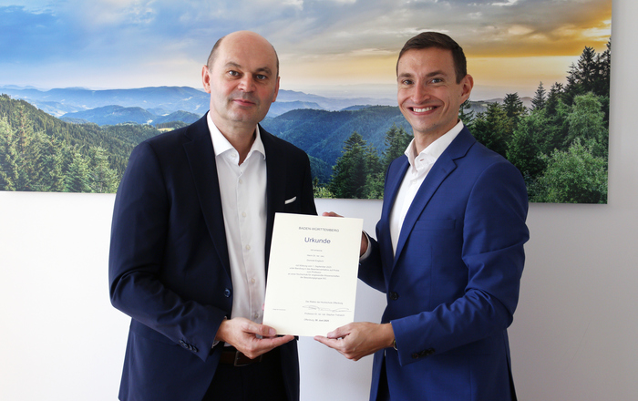 Zwei Männer im Anzug stehen vor einem Schwarzwaldfoto und halten eine Urkunde zwischen Sich