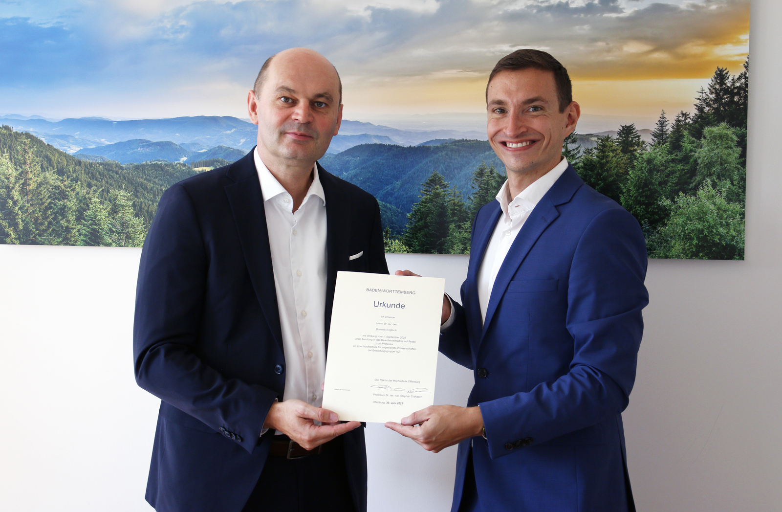Zwei Männer im Anzug stehen vor einem Schwarzwaldfoto und halten eine Urkunde zwischen Sich