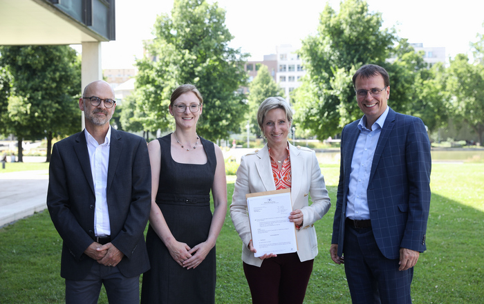 Prof. Dr. Tobias Hagen, Prof.in Dr. Simone Braun, Ministerin Dr. Nicole Hoffmeister-Kraut und Kultus-Staatssekretär Volker Schebesta (von links) stehen auf dem Rasen vor dem Ministerium. Die Ministerin hat den Bewilligungsbescheid in den Händen