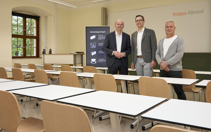 Drei Männer stehen in einem Raum mit Tischen und Stühlen