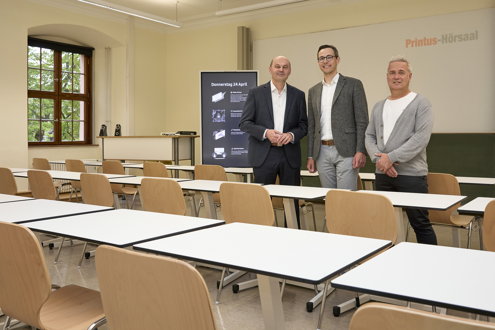 Drei Männer stehen in einem Raum mit Tischen und Stühlen