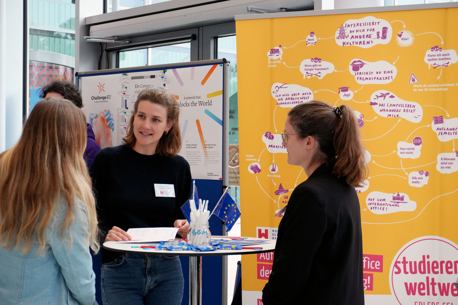 Drei Frauen stehen um einen Stehtisch herum und unterhalten sich im Hintergund sind Roll-ups zu sehen