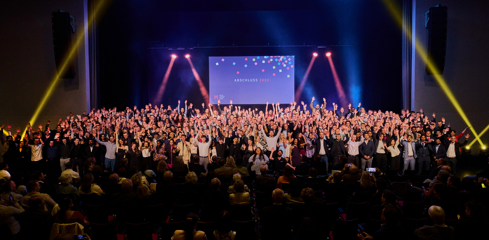 Gruppenfoto aller Absolventinnen und Absolventen mit jubelnd erhobenen Armen auf der Bühne auf einem Bildschirm über ihren Köpfen steht Abschluss 2025