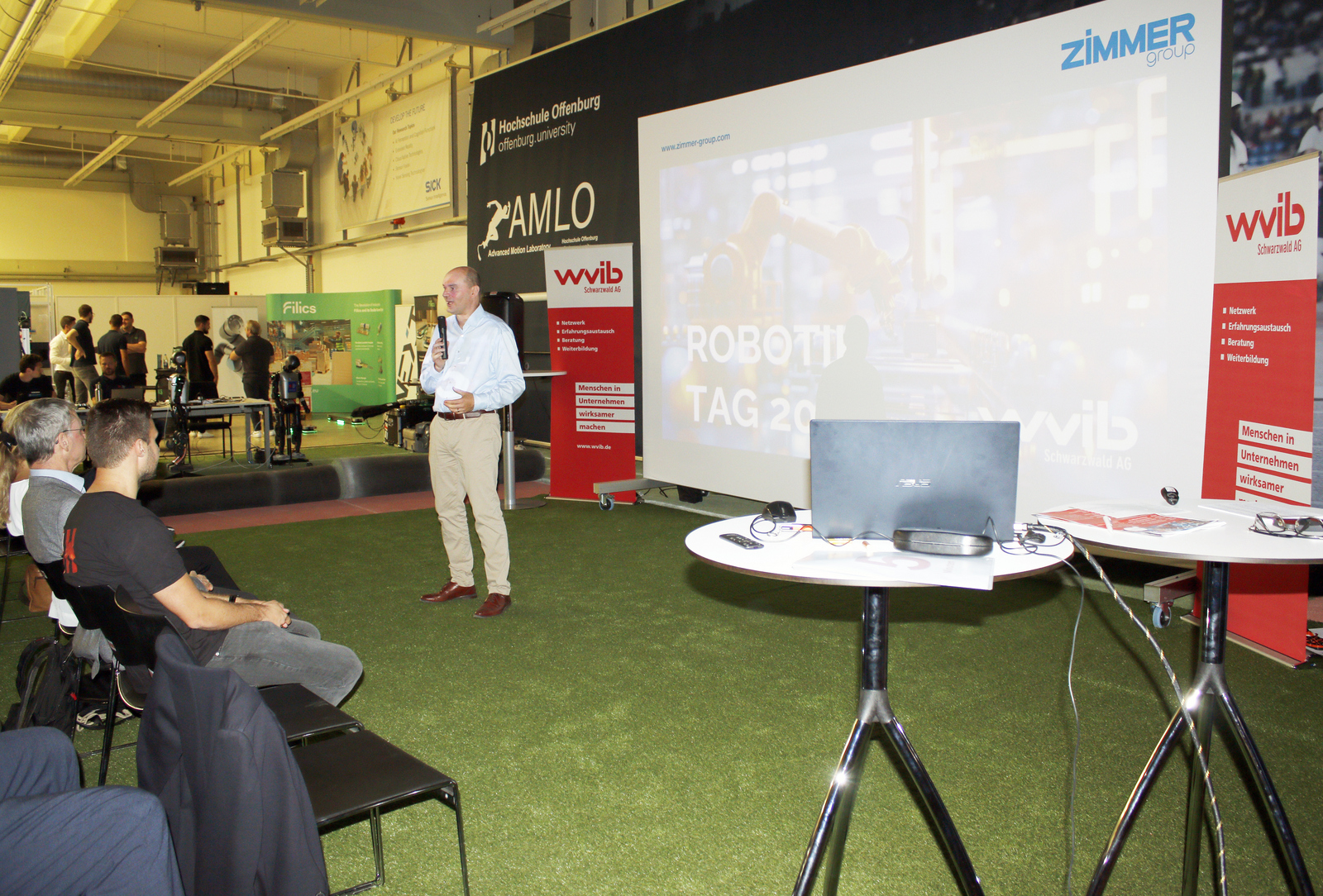 Ein Mann steht vor einer Leinwand vor sitzenden Zuhörenden und spricht ins Mikrofon