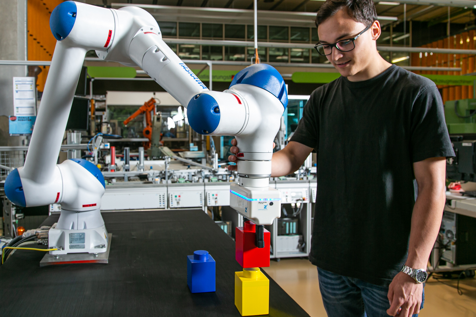 Ein junger Mann mit schwarzem T-Shirt und schwarzer Hose steht in einem Labor an einem Roboterarm der bunte Bauklötze stapelt