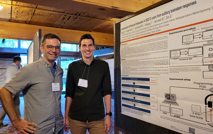 Stefan Zirn und Sebastian Roth stehen während der Konferenz neben dem Poster des akademischen Mitarbeiters.