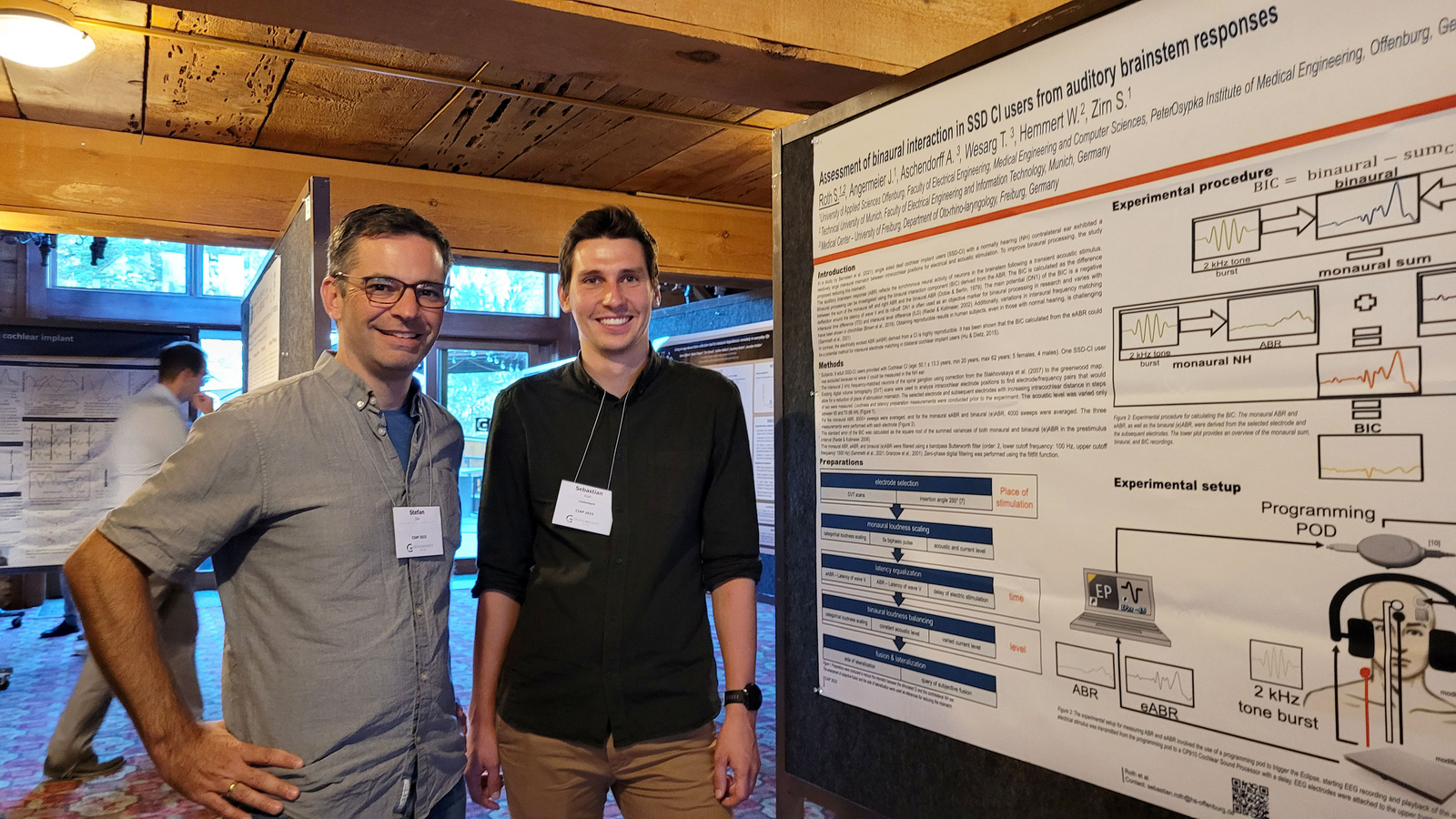 Stefan Zirn und Sebastian Roth stehen während der Konferenz neben dem Poster des akademischen Mitarbeiters.