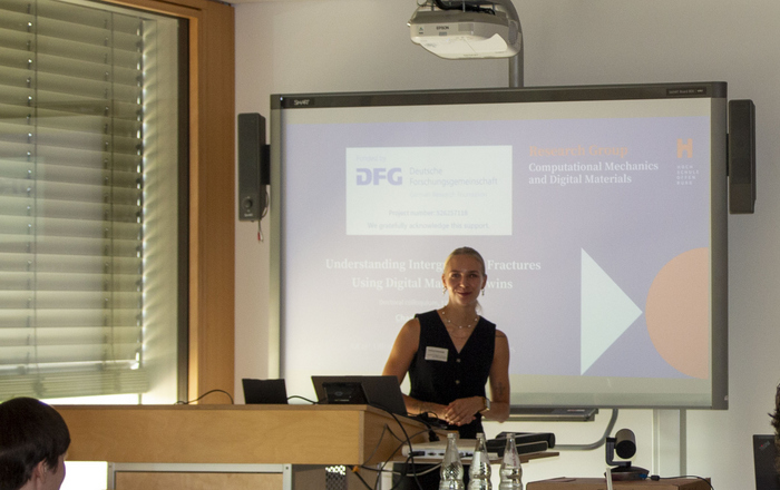 Eine junge Frau steht in einem Hörsaal vor einem Bildschirm auf dem eine Präsentation zu sehen ist