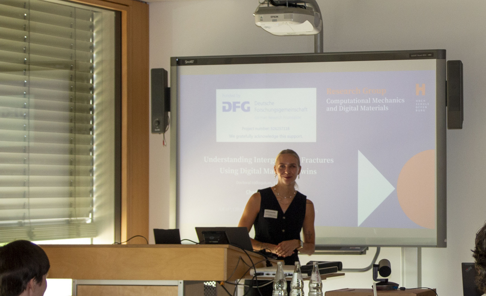 Eine junge Frau steht in einem Hörsaal vor einem Bildschirm auf dem eine Präsentation zu sehen ist