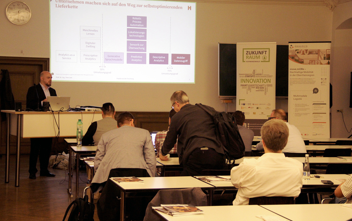 Ein Mann steht vor sitzenden Zuhörenden neben einer Leinwand am Rednerpult