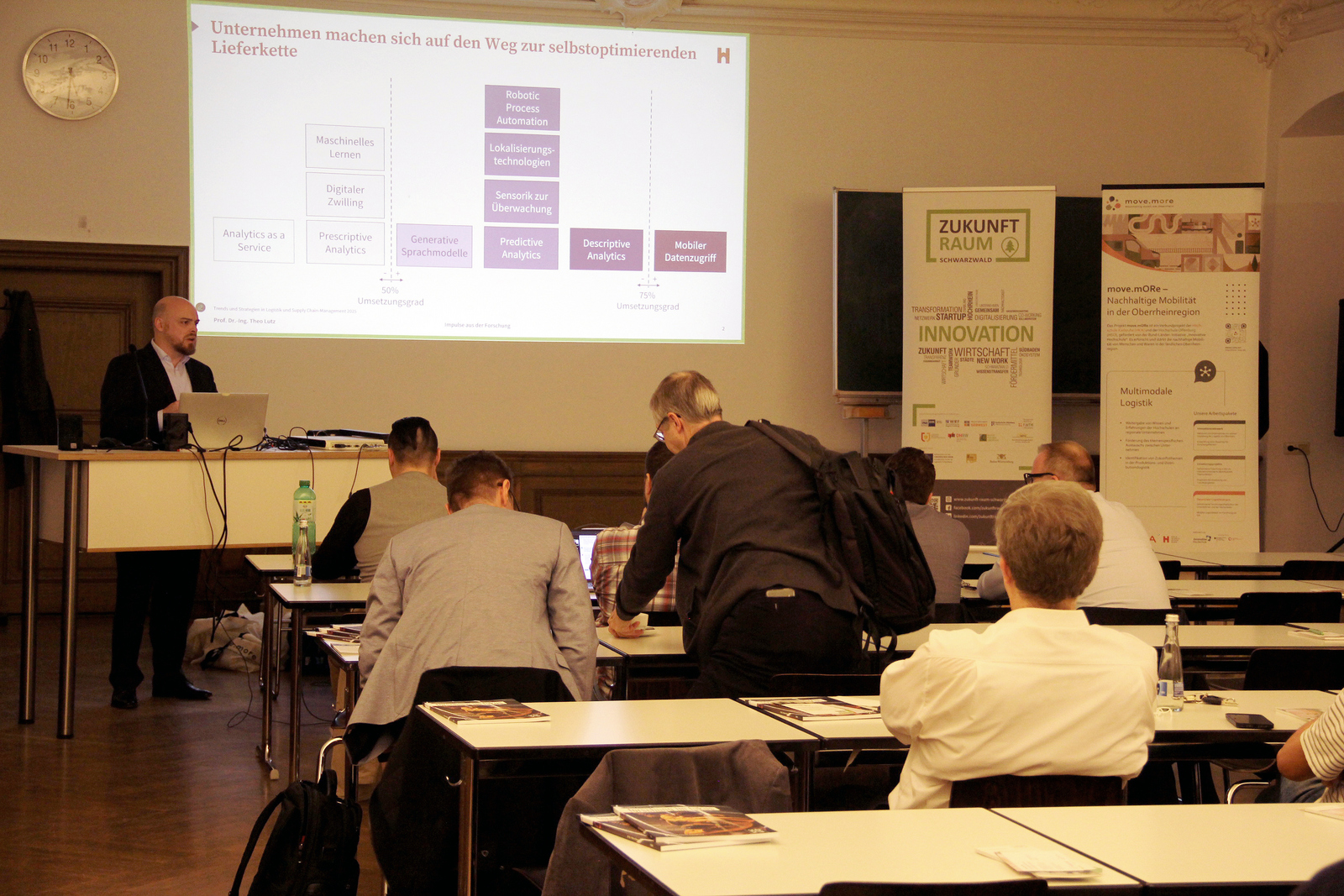 Ein Mann steht vor sitzenden Zuhörenden neben einer Leinwand am Rednerpult