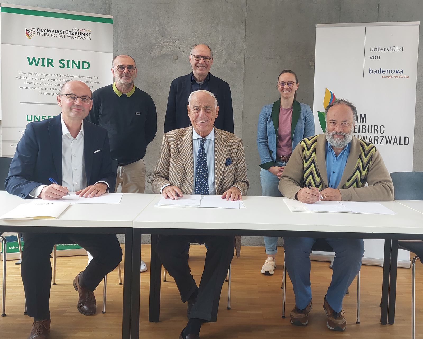 Drei Männer sitzen nebeneinander an einem Tisch und unterzeichnen die Kooperationsvereinbarung. Drei Menschen stehen hinter ihnen.