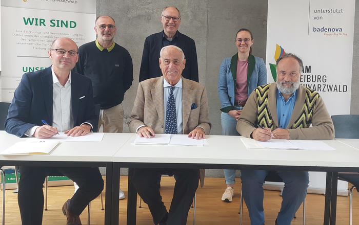 Drei Männer sitzen nebeneinander an einem Tisch und unterzeichnen die Kooperationsvereinbarung. Drei Menschen stehen hinter ihnen.