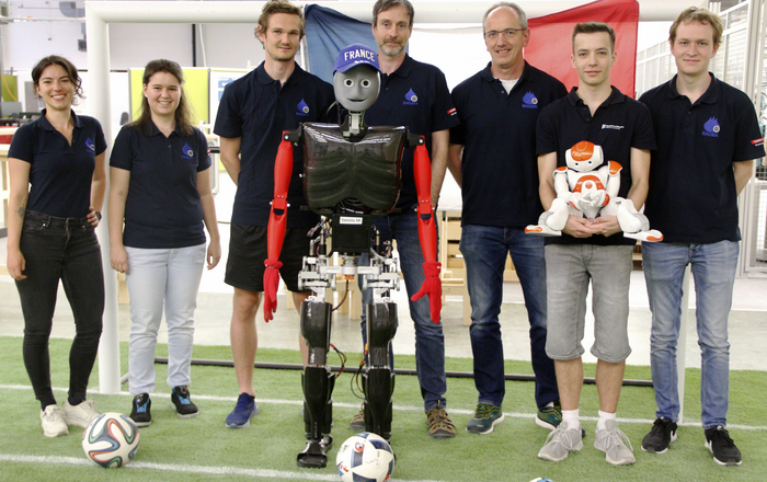 Gruppenfoto mit Robotern vor einem Tor in dem eine Frankreichflagge hängt, Sweaty hat eine blaue Baseball-Kappe mit dem weißen Schriftzug France auf.