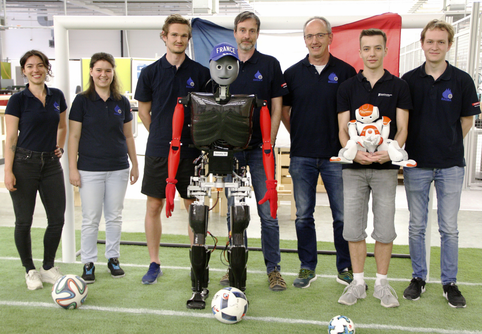 Gruppenfoto mit Robotern vor einem Tor in dem eine Frankreichflagge hängt, Sweaty hat eine blaue Baseball-Kappe mit dem weißen Schriftzug France auf.