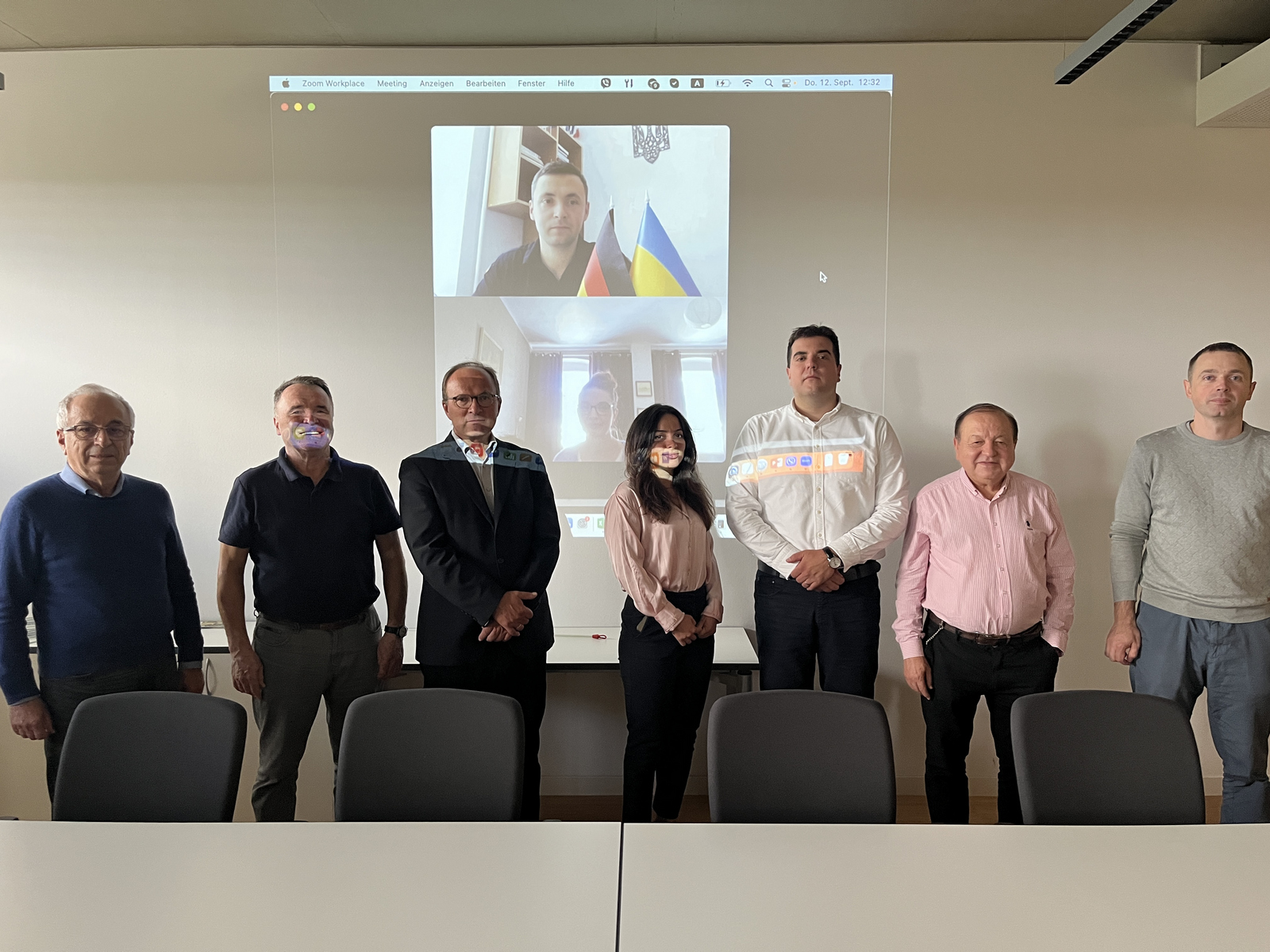 7 Personen in einer Reihe vor der Leinwand, auf der der 8. Teilnehmer zu sehen ist