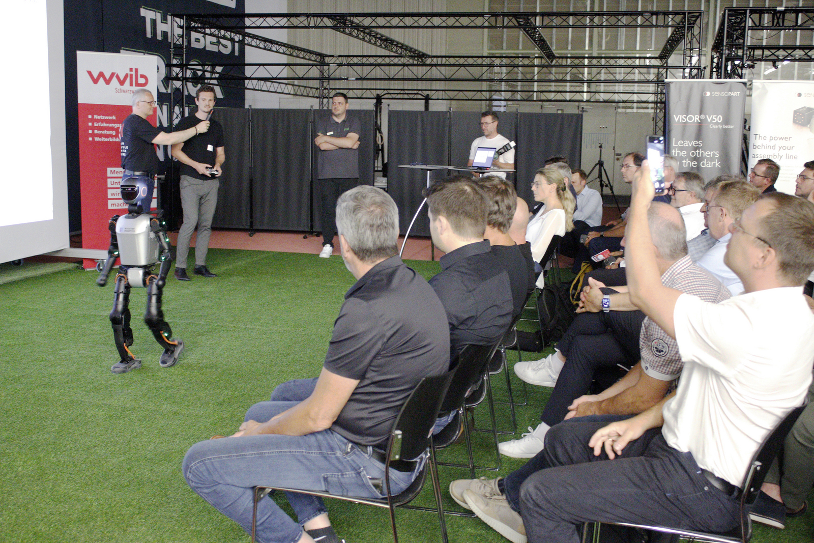 Ein Mann steht vor sitzenden Zuhördenden und führt einen humanoiden Roboter vor