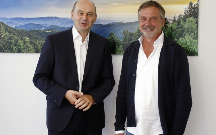 Zwei Männer stehen vor einem Schwarzwaldfoto und schauen in die Kamera