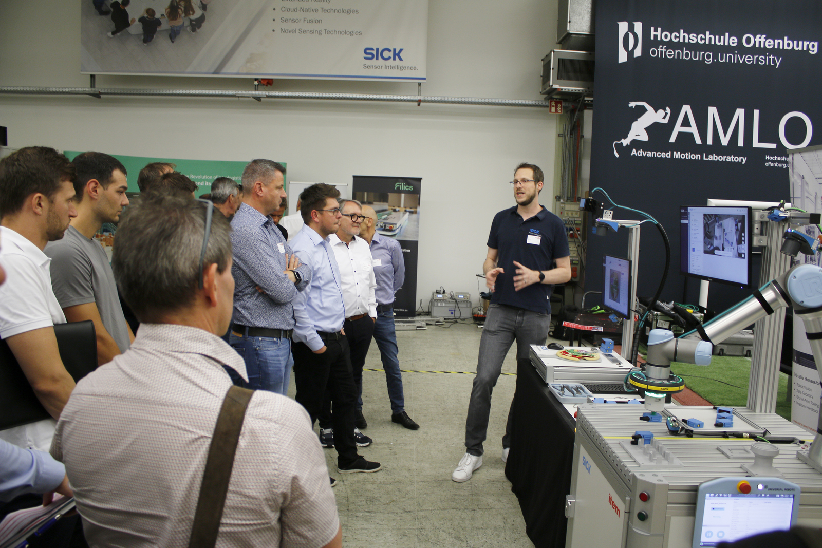 Ein Mann steht neben zwei kleinen Industrierobotern und erklärt den Umstehenden etwas