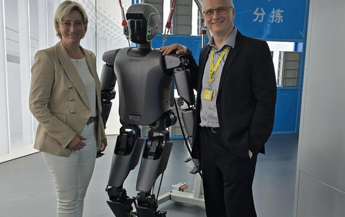 Eine Frau und ein Mann stehen links und rechts von einem humanoiden Roboter und legen diesem jeweils eine Hand auf die Schultern