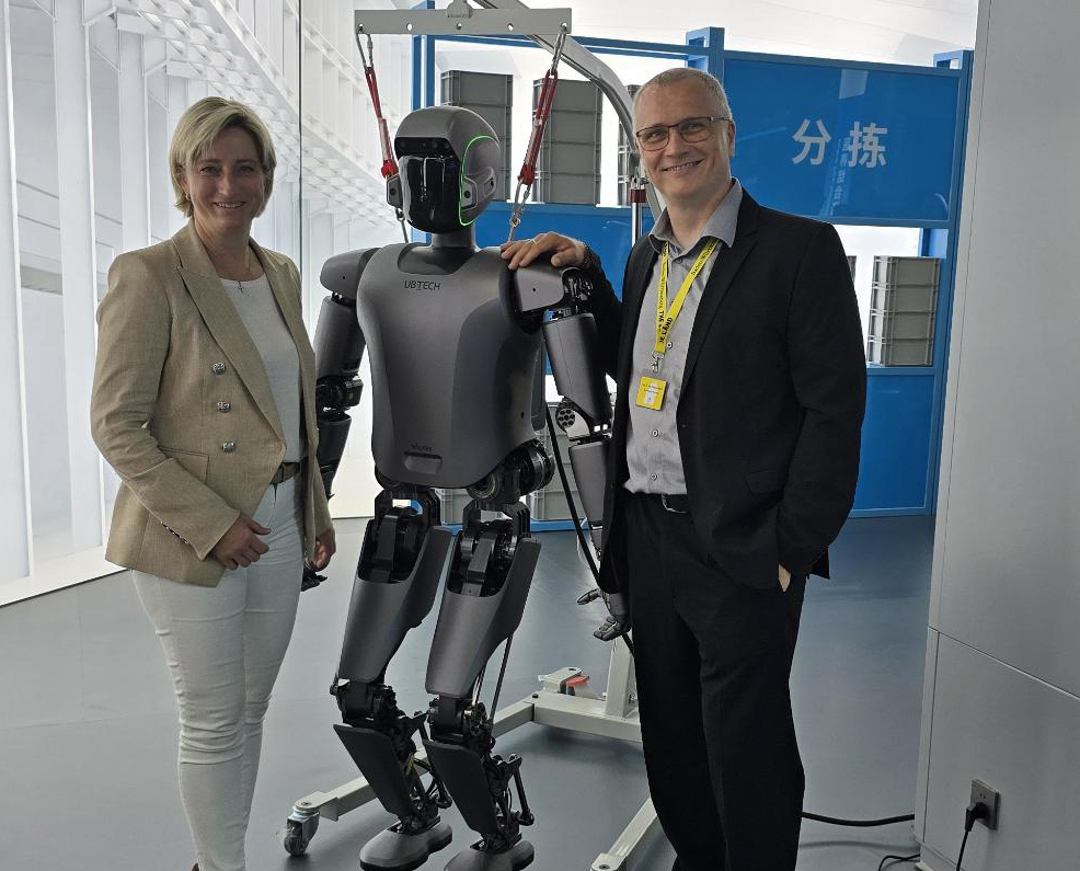 Eine Frau und ein Mann stehen links und rechts von einem humanoiden Roboter und legen diesem jeweils eine Hand auf die Schultern