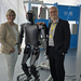 Eine Frau und ein Mann stehen links und rechts von einem humanoiden Roboter und legen diesem jeweils eine Hand auf die Schultern