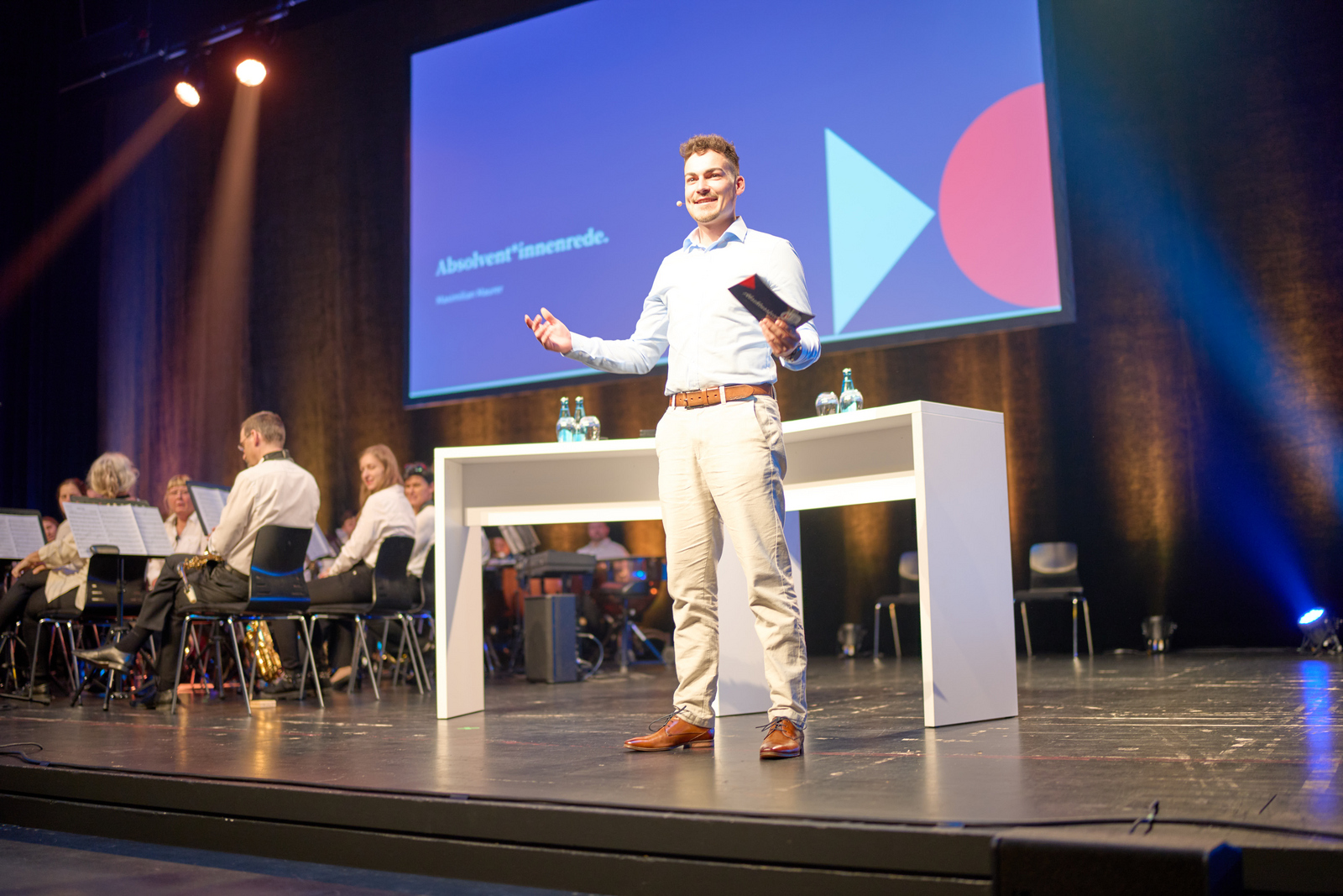 Ein junger Mann in heller Hose und Hemd steht auf der Bühne vor einem Tisch und hält eine Rede