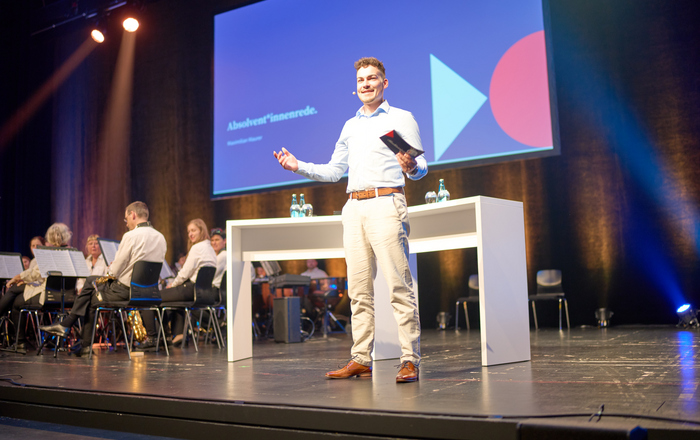 Ein junger Mann in heller Hose und Hemd steht auf der Bühne vor einem Tisch und hält eine Rede