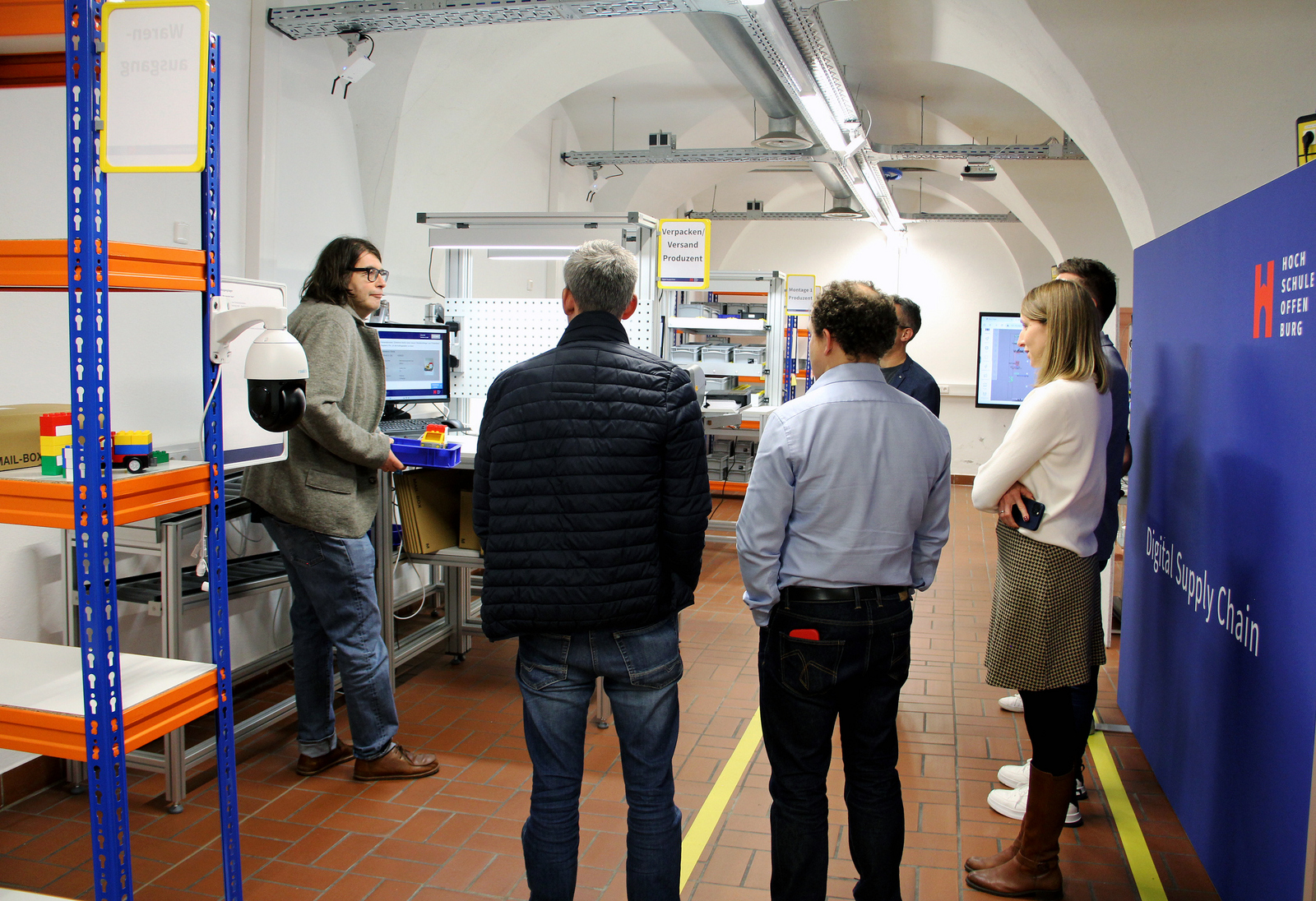 Ein Mann macht in einem Labor an einem Tisch mit Bildschirm Waren versandfertig währen eine Gruppe Menschen um ihn herum steht