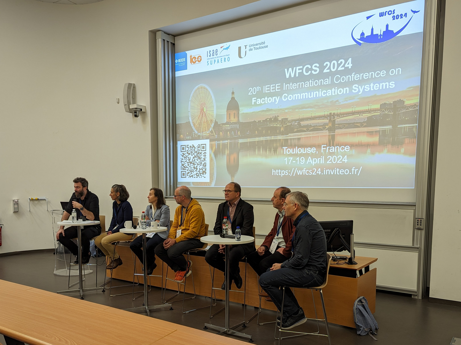 sieben Personen sitzen in einer Reihe vor einer Päsentationswand