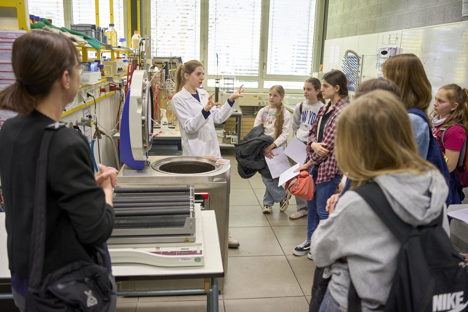 Mädchen stehen in einem LAbor vor einer Zentrifuge eine Frau im weißen Kittel erklärt etwas