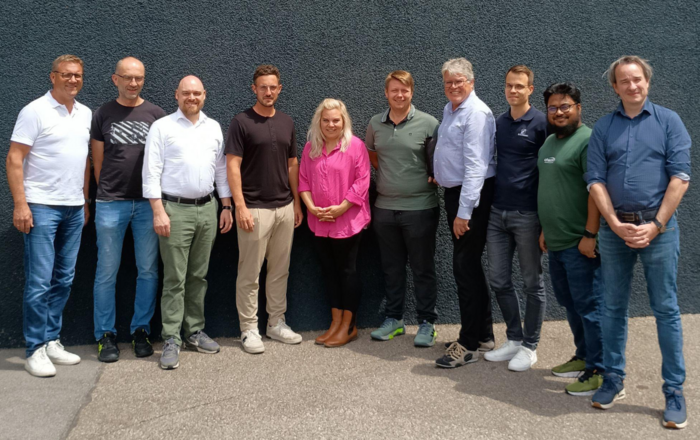 Gruppe von elf Personen steht nebeneinander vor einer grauen Wand