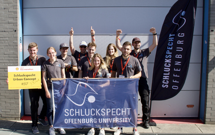 Das jubelnde Team mit Teamfahnen und einem Startschild auf dem handschriftlich die Kilometerleistung mit einem Liter Diesel notiert ist vor einem Garagentor 