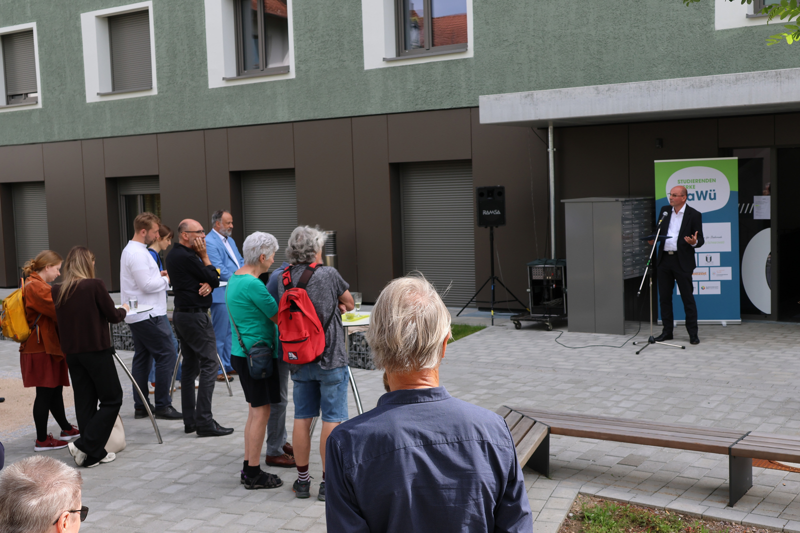 Ein Mann steht vor einem Gebäude und spricht zu Menschen an Stehtischen