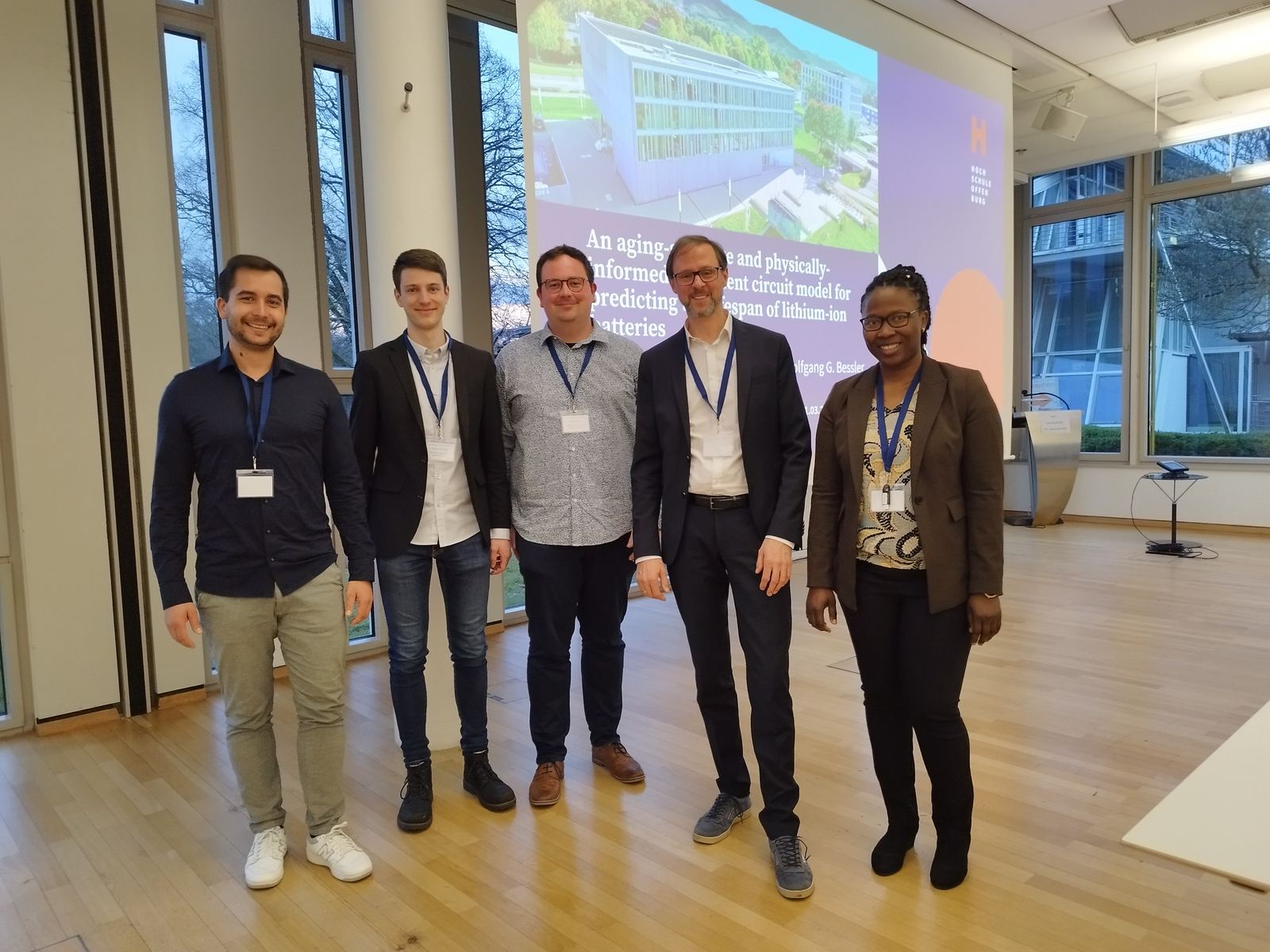 fünf Menschen stehen vor einer Leinwand auf der eine Präsentation zu sehen ist und schauen in die Kamera