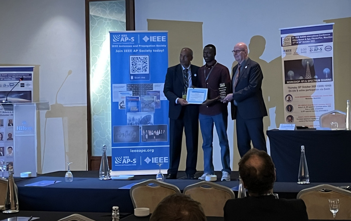 Drei Männer stehe zwischen Roll-ups auf einer Bühne der Mittlere erhält eine Urkunde