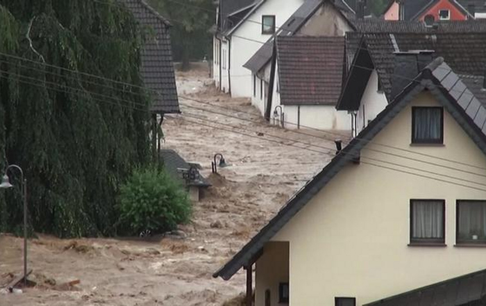 Eine braune Flutwelle bahnt sich ihren Weg zwischen Häusern und Bäumen hindurch 