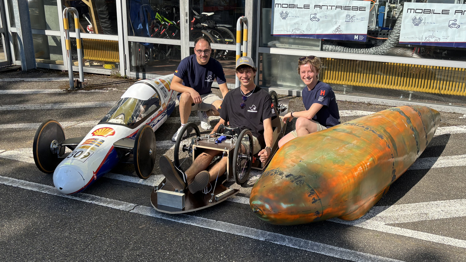 Drei jungen Menschen zwischen beziehungsweise einem Rennwagen, einem Versuchträger für das neue Fahrwerk sowie einer 3D-gedruckten Karosserie. 