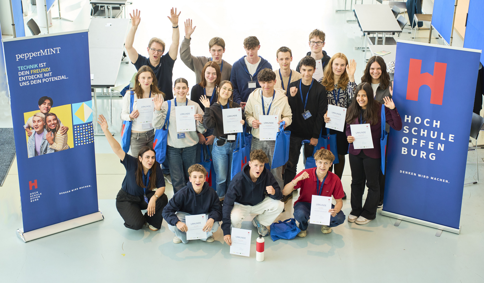 Gruppenfoto der zwischen zwei Roll-ups knieenden und stehenden Teilnehmenden mit Urkunden