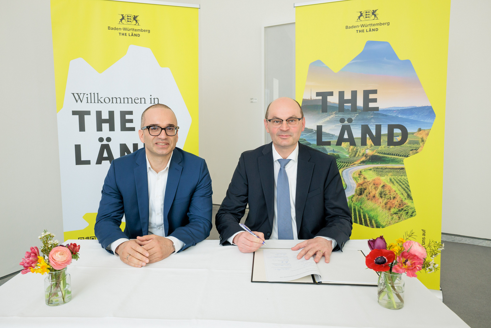 Zwei Männer sitzen vor gelben The Länd-Roll-ups an einem Tisch und unterzeichnen die Vereinbarung