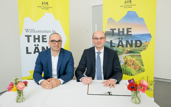 Zwei Männer sitzen vor gelben The Länd-Roll-ups an einem Tisch und unterzeichnen die Vereinbarung