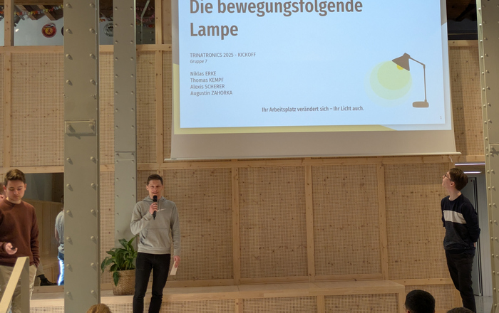 Junge Männer erklären in einer hohen Halle eine Präsentation auf einer Leinwand