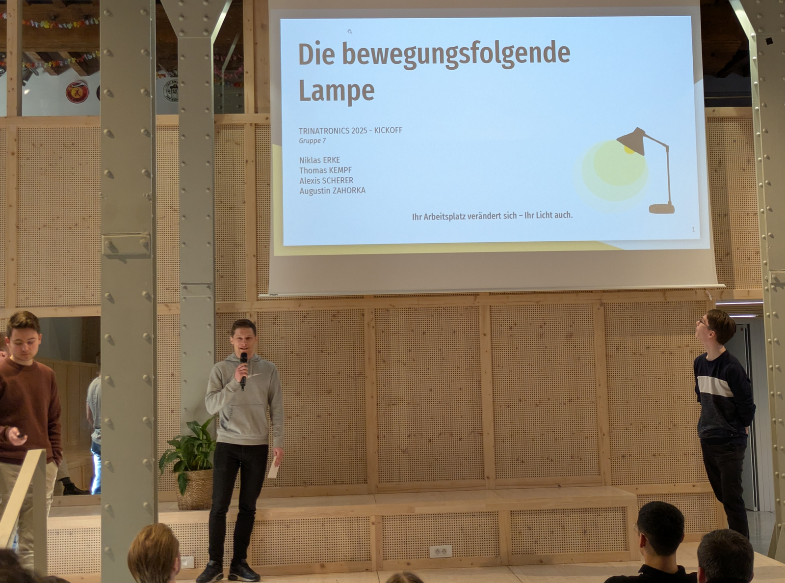 Junge Männer erklären in einer hohen Halle eine Präsentation auf einer Leinwand