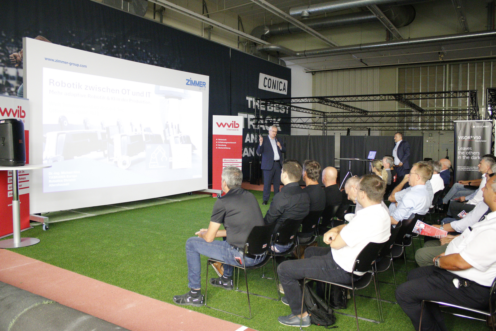 Ein Mann steht neben einer Leinwand und hält einen Vortrag für die sitzenden Zuhörenden 