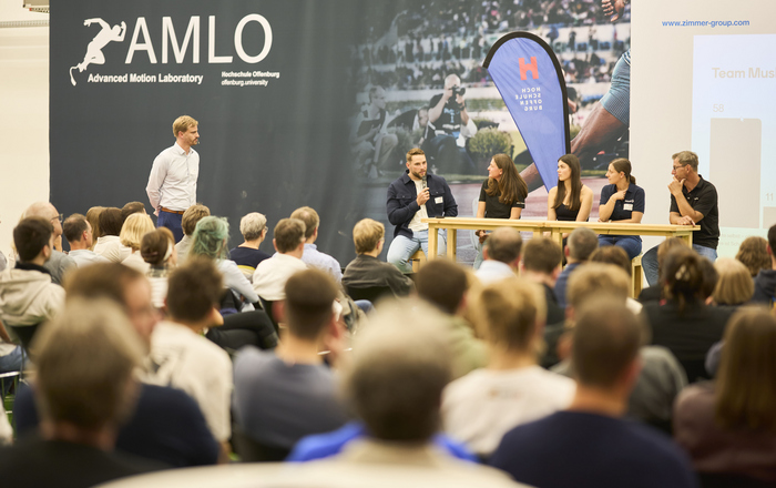 Blick über sitzende Menschen hinweg auf die Bühne wo vier Personen sitzen ein Mann steht links von ihnen