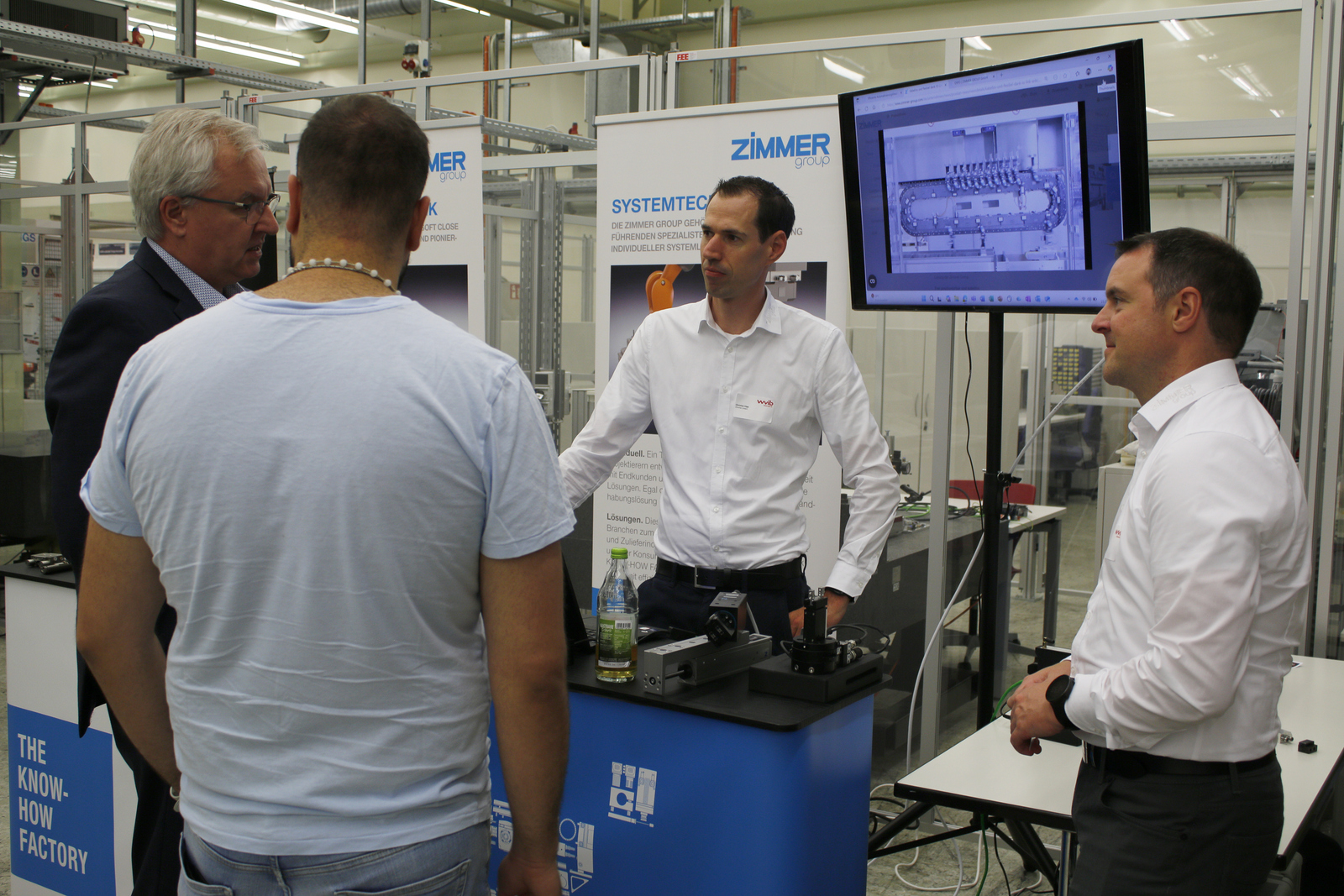 Ein Mann steht hinter einem Messestand und erklärt den Davorstehenden was auf dem Bildschirm rechts neben ihm zu sehen ist