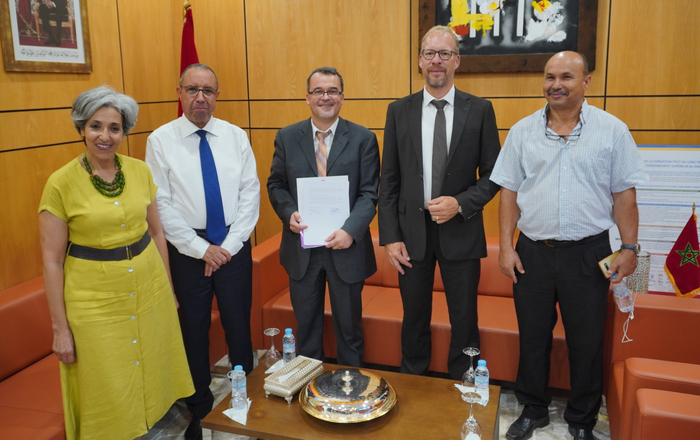 Die Personen stehen an einem kleinen Couchtisch in einem Büro und die mittlere Person hält das Abkommen in den Händen.