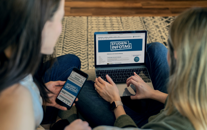 Zwei Mädchen sitzen auf dem Boden vor einem aufgeklappten Laptop auf dem Studienfotag zu lesen ist.